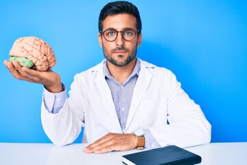Young hispanic man sitting at the table wearing doctor coat holding brain thinking attitude and...