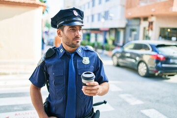 Young hispanic policeman wearing police uniform with serious expression. Drinking cup of take away coffee standing at town street.