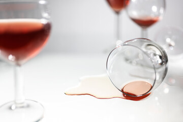 Overturned drinking glass with a puddle of red wine between blurred standing glasses, concept for party fun, alcohol addiction or burn out, light gray background with copy space, selected focus