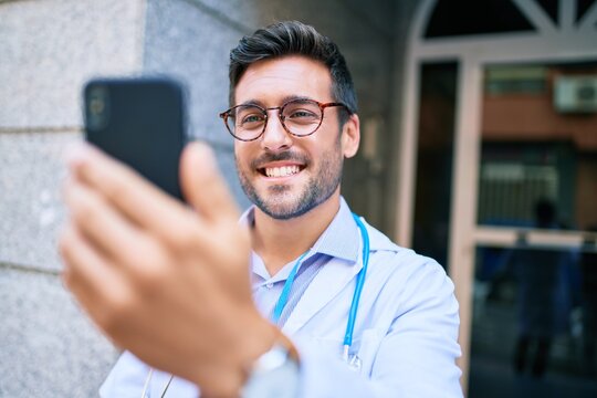 Young handsome hispanic doctor wearing uniform and stethoscope smiling happy Standing with smile on face using smartphone at town street.