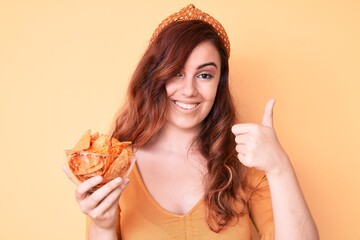Young beautiful woman holding nachos potato chips smiling happy and positive, thumb up doing...