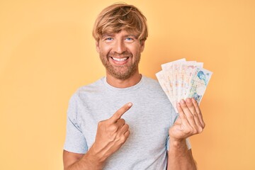 Young blond man holding united kingdom pounds smiling happy pointing with hand and finger