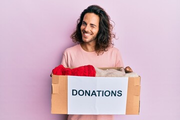 Young handsome man with long hair holding donations box for charity winking looking at the camera...
