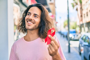 Young hispanic man smiling happy holding hiv awareness red ribbon at city.