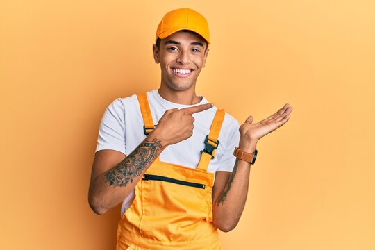 Young handsome african american man wearing handyman uniform over yellow background amazed and smiling to the camera while presenting with hand and pointing with finger.