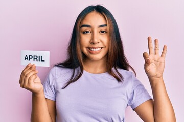 Young latin woman holding april word paper doing ok sign with fingers, smiling friendly gesturing...