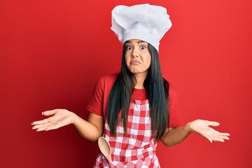 Young beautiful hispanic girl wearing baker uniform and cook hat clueless and confused expression with arms and hands raised. doubt concept.