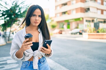 Young hispanic girl with serious expression using smartphone and drinking take away coffee at the city.