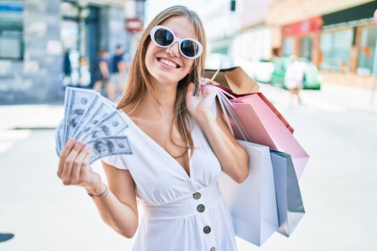 Young blonde woman smiling happy holding shopping bags and dollars banknotes at street of city