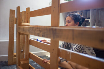 Young Attractive Hispanic Woman in Black and White Stripes Long Sleeve Shirt and Black Shorts Paints a Wooden Pallet in White Smiling at Home