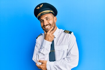 Handsome man with beard wearing airplane pilot uniform looking confident at the camera with smile with crossed arms and hand raised on chin. thinking positive.