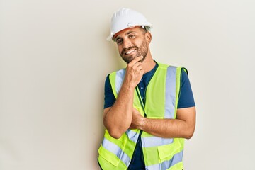 Handsome man with beard wearing safety helmet and reflective jacket smiling looking confident at the camera with crossed arms and hand on chin. thinking positive.