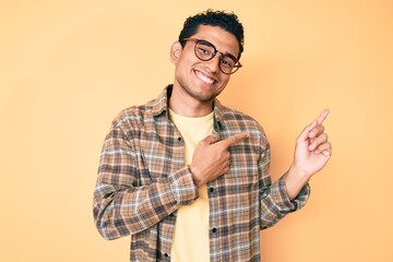 Young handsome hispanic man wearing casual clothes and glasses smiling and looking at the camera pointing with two hands and fingers to the side.