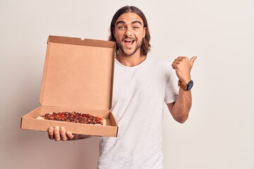 Young handsome man holding delivery pizza box pointing thumb up to the side smiling happy with open mouth