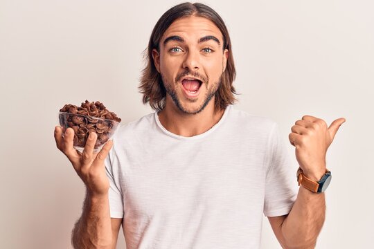 Young handsome man holding chocolate cereals pointing thumb up to the side smiling happy with open mouth