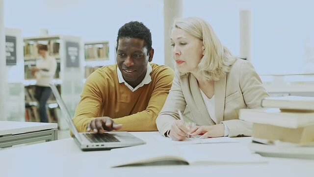 Friendly female tutor helping to diligent african-american man preparing for exam in library