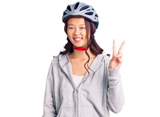 Young beautiful chinese girl wearing bike helmet smiling with happy face winking at the camera doing victory sign. number two.