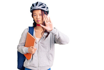 Young beautiful chinese girl wearing student backpack and bike helmet holding book with open hand doing stop sign with serious and confident expression, defense gesture