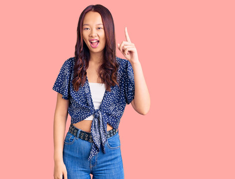 Young beautiful chinese girl wearing casual clothes pointing finger up with successful idea. exited and happy. number one.