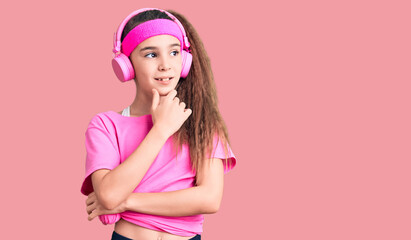 Obraz na płótnie Canvas Cute hispanic child girl wearing gym clothes and using headphones with hand on chin thinking about question, pensive expression. smiling with thoughtful face. doubt concept.