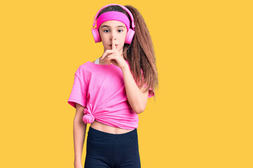 Cute hispanic child girl wearing gym clothes and using headphones asking to be quiet with finger on...