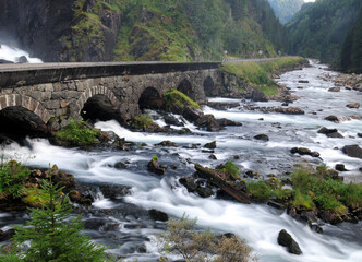 Fototapeta na wymiar Long Time Exposure Of Latefossen Falls Flowing Into The Gronsdalslona River Passing A Stone Bridge On A Cloudy Summer Day