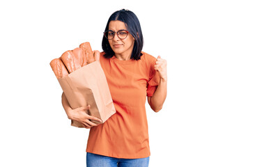 Obraz na płótnie Canvas Young beautiful latin woman wearing glasses holding paper bag with bread annoyed and frustrated shouting with anger, yelling crazy with anger and hand raised