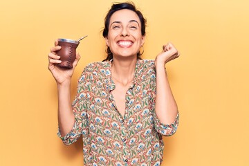 Young beautiful hispanic woman drinking mate infusion screaming proud, celebrating victory and...