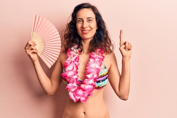 Young beautiful hispanic woman wearing bikini and hawaiian lei holding hand fan smiling with an...