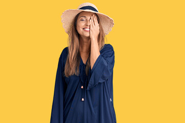 Middle age hispanic woman wearing summer hat covering one eye with hand, confident smile on face and surprise emotion.
