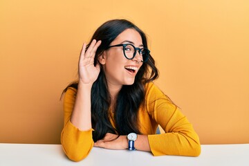 Beautiful brunette young woman wearing glasses and casual clothes sitting on the table smiling with...