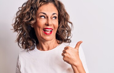 Middle age beautiful brunette woman wearing casual t-shirt standing over white background pointing thumb up to the side smiling happy with open mouth