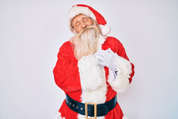 Old senior man with grey hair and long beard wearing santa claus costume with suspenders smiling...