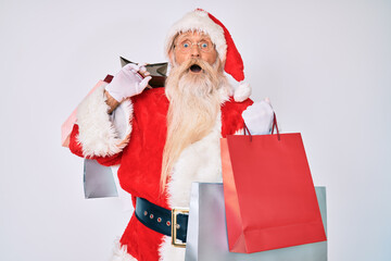 Old senior man with grey hair and long beard wearing santa claus costume holding shopping bags afraid and shocked with surprise and amazed expression, fear and excited face.