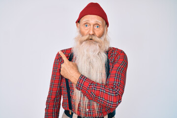 Old senior man with grey hair and long beard wearing hipster look with wool cap surprised pointing with finger to the side, open mouth amazed expression.