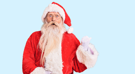 Old senior man with grey hair and long beard wearing traditional santa claus costume surprised pointing with hand finger to the side, open mouth amazed expression.
