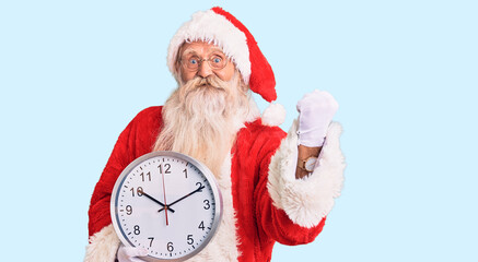 Old senior man with grey hair and long beard wearing santa claus costume holding clock screaming proud, celebrating victory and success very excited with raised arms