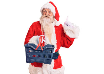 Old senior man with grey hair and long beard wearing santa claus costume holding shopping basket surprised with an idea or question pointing finger with happy face, number one