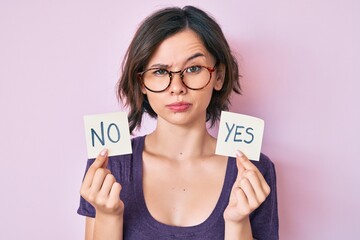 Young beautiful girl holding yes and no reminder skeptic and nervous, frowning upset because of...