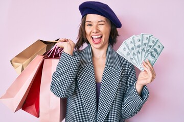 Young beautiful woman wearing beret holding shopping bags and dollars banknotes winking looking at...