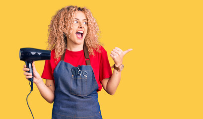 Young blonde woman with curly hair wearing hairdresser apron and holding dryer blow pointing thumb...