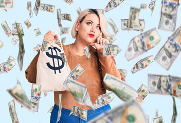 Young blonde plus size woman holding money bag with dollar symbol serious face thinking about...