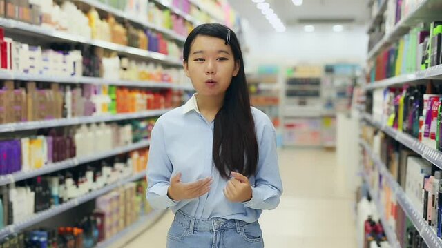 Portrait of friendly Asian female shop assistant near shelves with hair dye in cosmetics shop