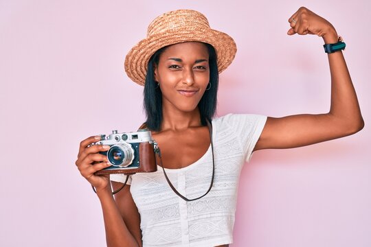 Young african american woman wearing summer hat holding vintage camera strong person showing arm muscle, confident and proud of power