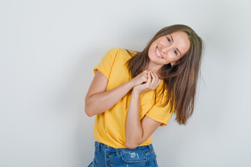 Young woman posing like asking for a favor in yellow t-shirt, shorts and looking cheerful , front view.