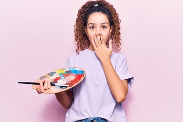 Beautiful kid girl with curly hair holding paintbrush and palette covering mouth with hand, shocked and afraid for mistake. surprised expression