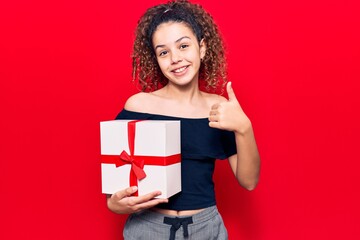 Beautiful kid girl with curly hair holding gift smiling happy and positive, thumb up doing excellent and approval sign