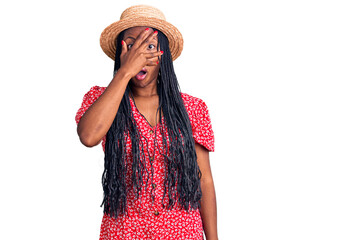 Young african american woman wearing summer hat peeking in shock covering face and eyes with hand, looking through fingers with embarrassed expression.