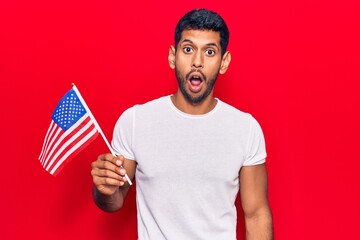 Young latin man holding united states flag scared and amazed with open mouth for surprise,...