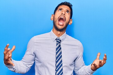 Young latin man wearing business clothes crazy and mad shouting and yelling with aggressive expression and arms raised. frustration concept.